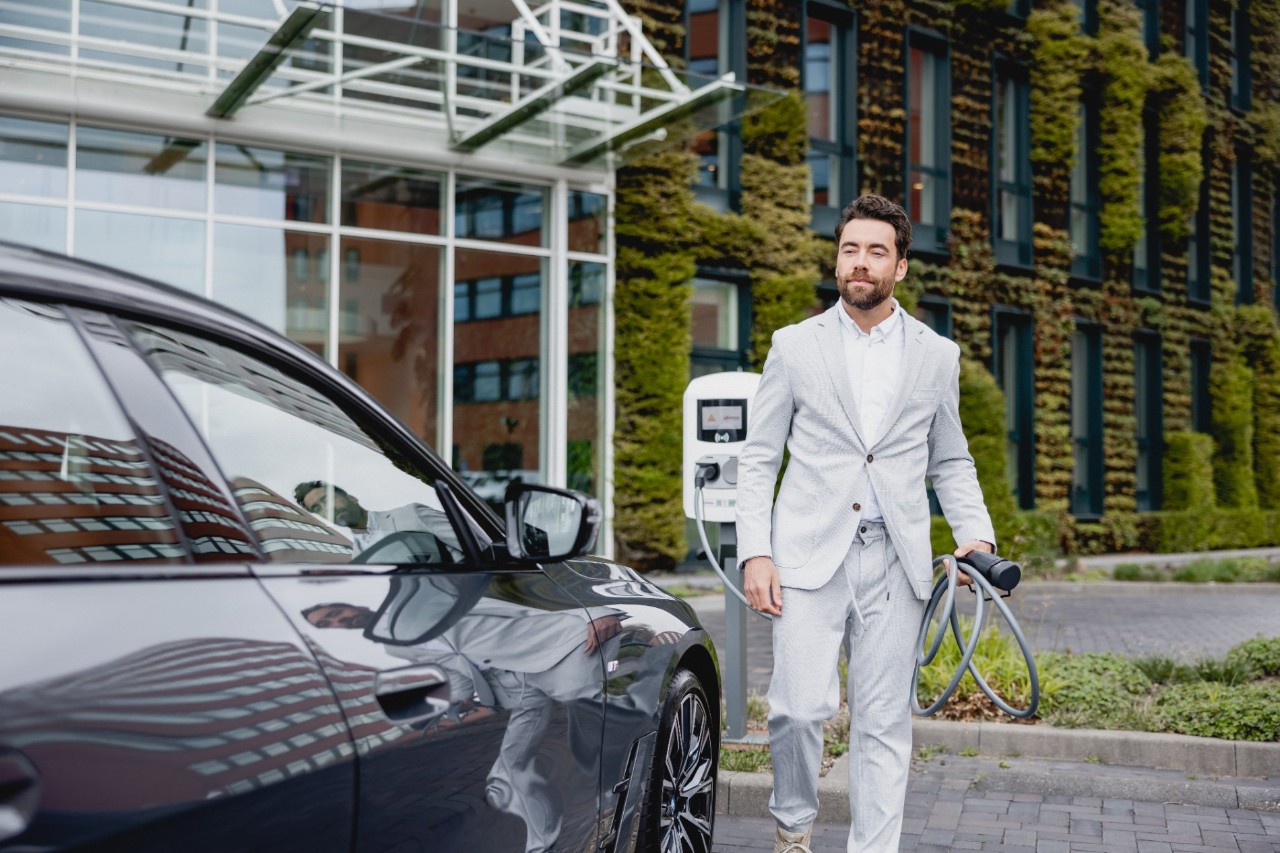 Man in a light suit stands next to an electric car and holds a charging cable at a charging station outside a modern building.