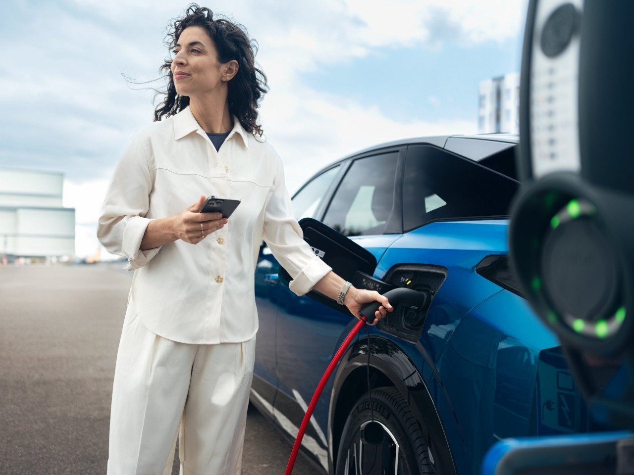 Woman holds a charging cable and smartphone next to a blue electric car at a charging station.
