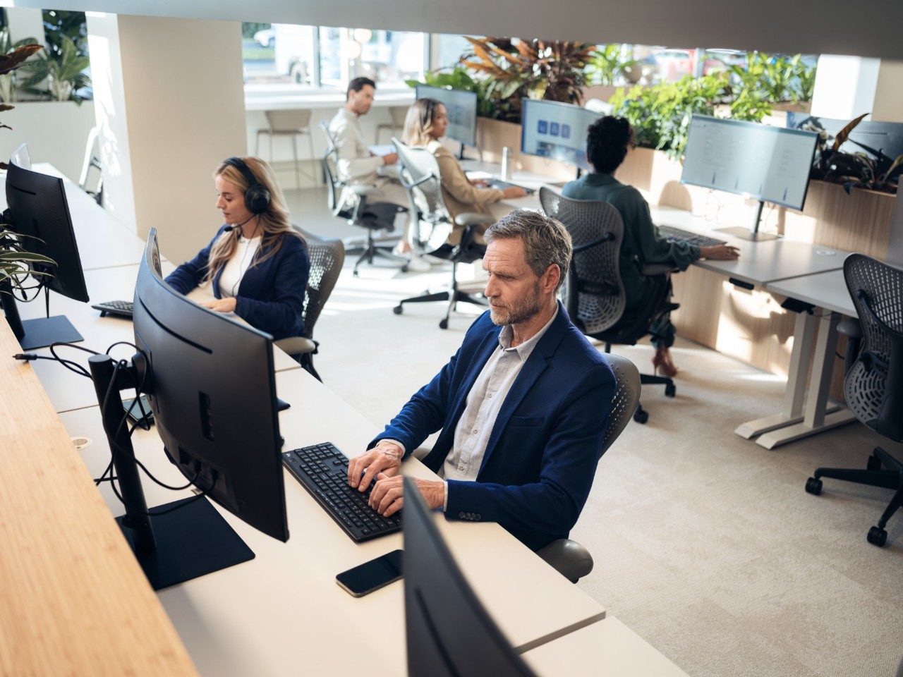 Open-plan modern office with employees working at computer workstations, surrounded by natural light and indoor plants.