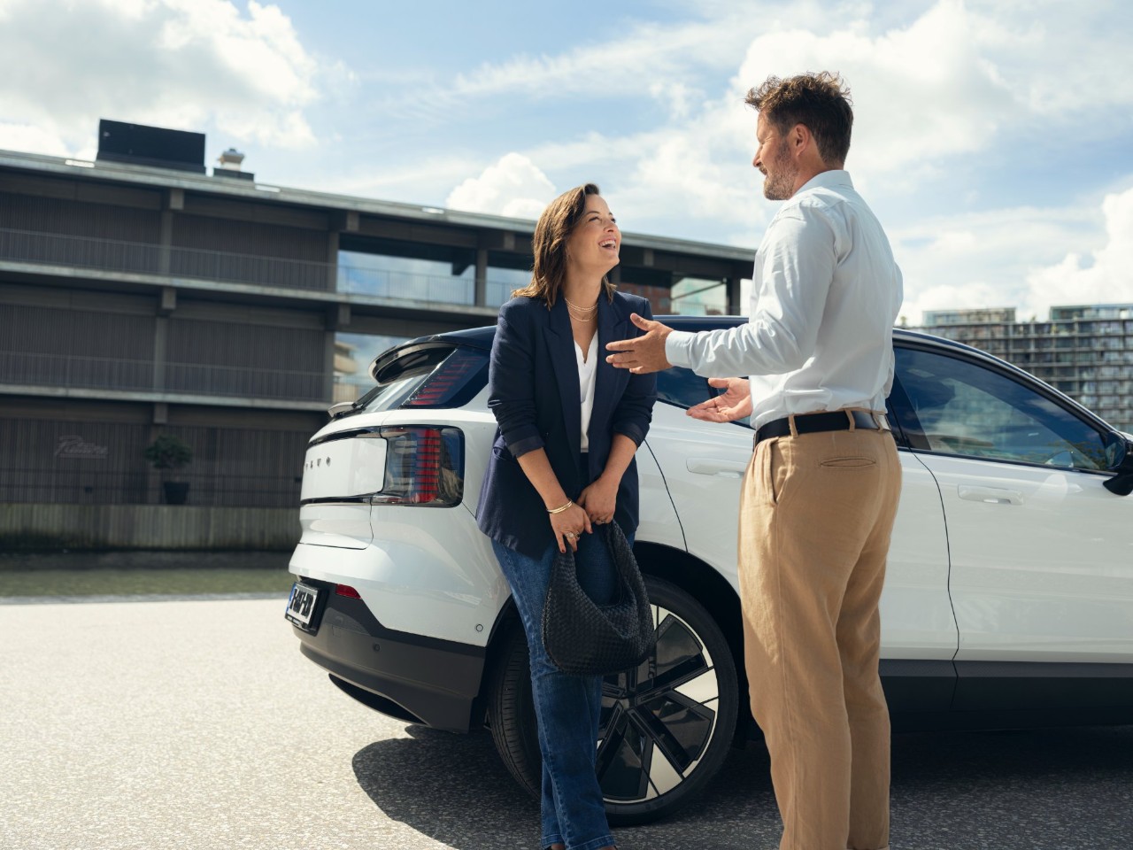 A woman leans against a white car and has a conversation with a man.