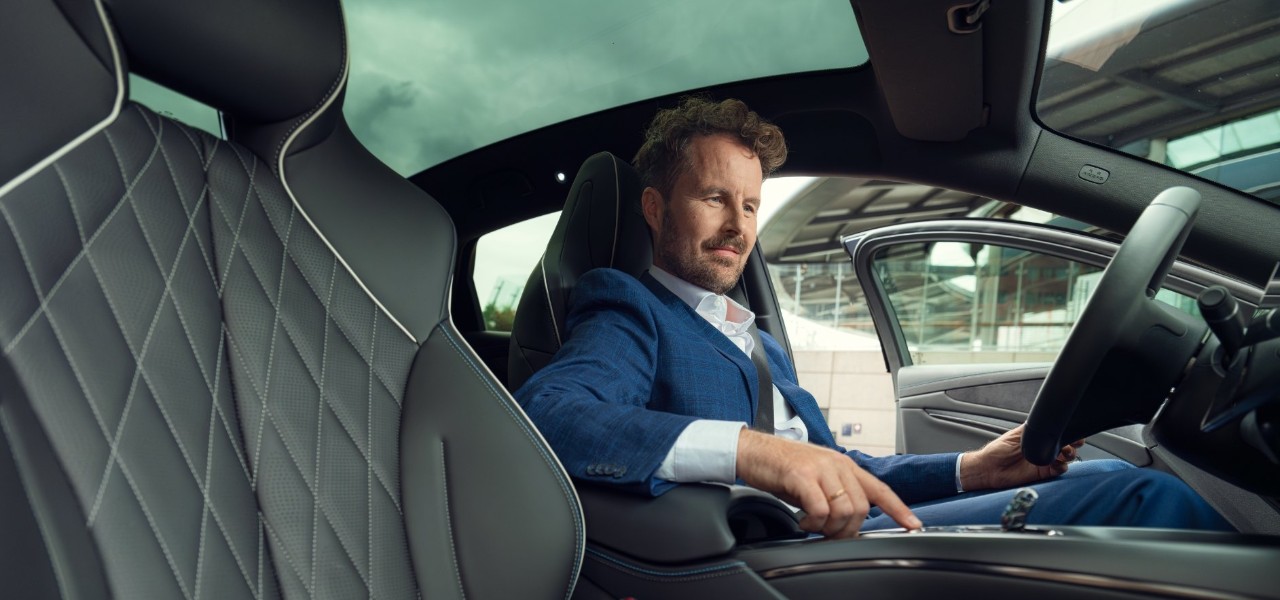 Person in a blue suit seated indoors near a modern structure, hand resting on an armrest.