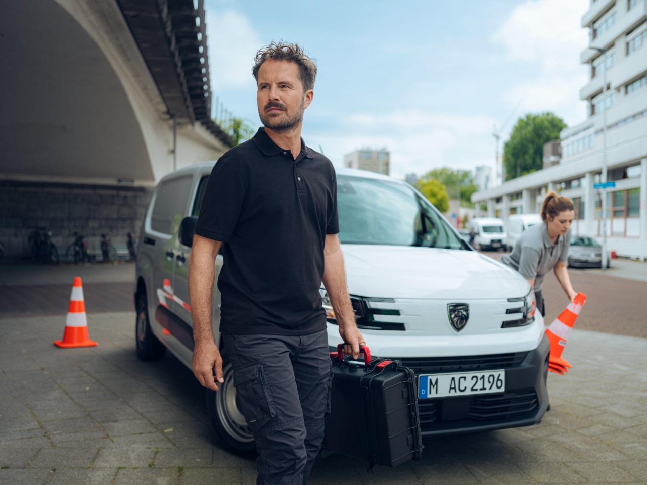 Man with a tool bag next to a delivery van in front of a bridge; woman with a traffic cone in the background.