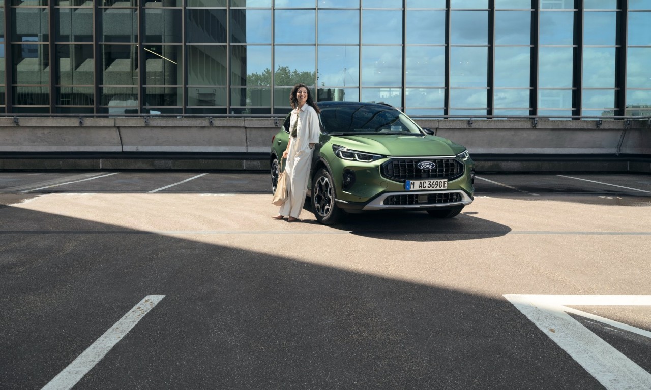 Woman leans against a green Ford vehicle in a parking lot in front of a glass facade.
