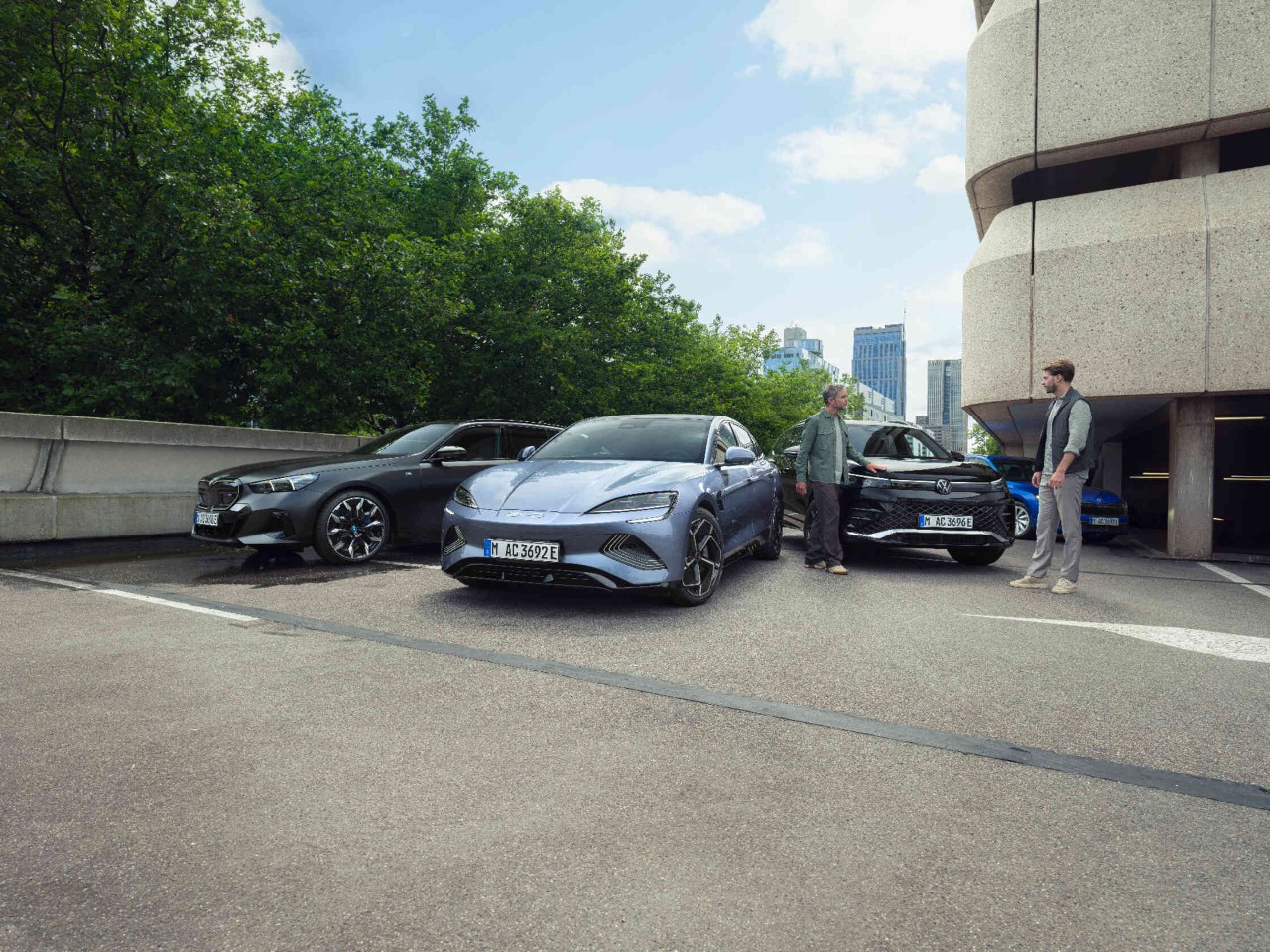Zwei Maenner neben drei Autos auf Parkplatz vor Parkdeck.