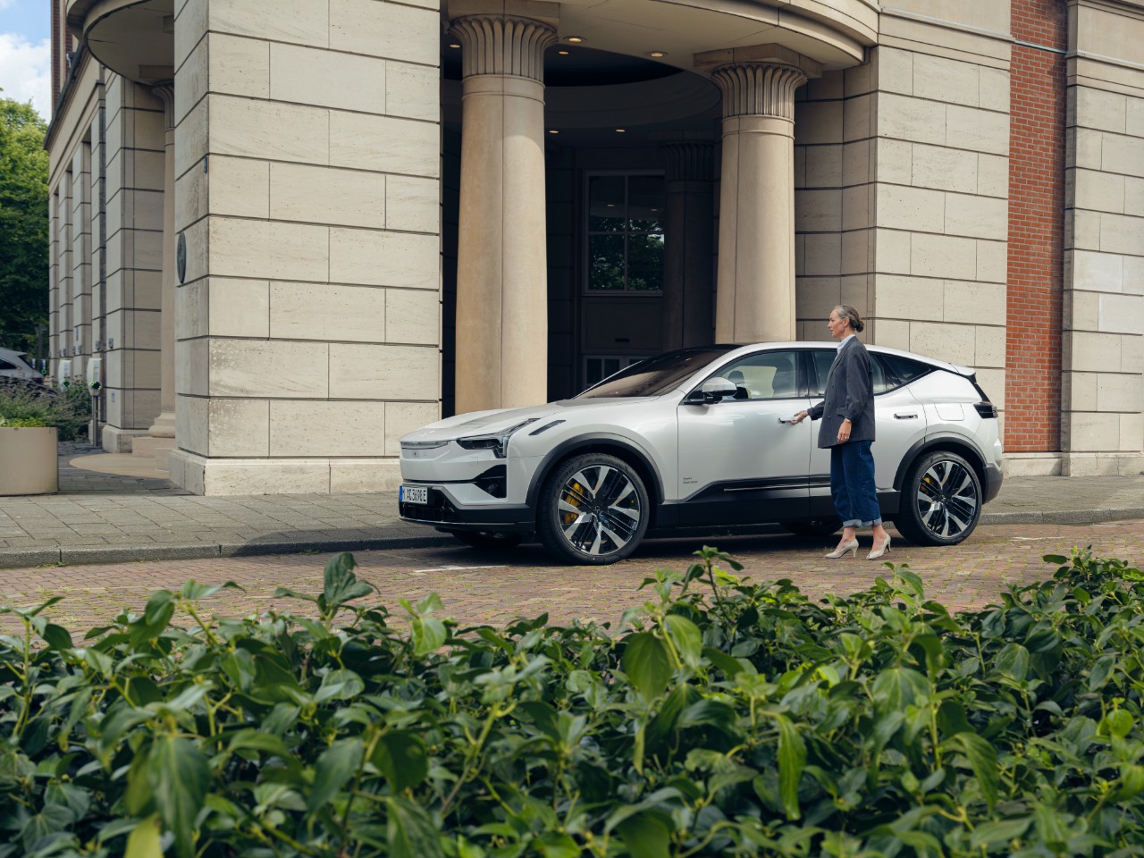 Woman stands in front of a silver car in front of an elegant building with columns.