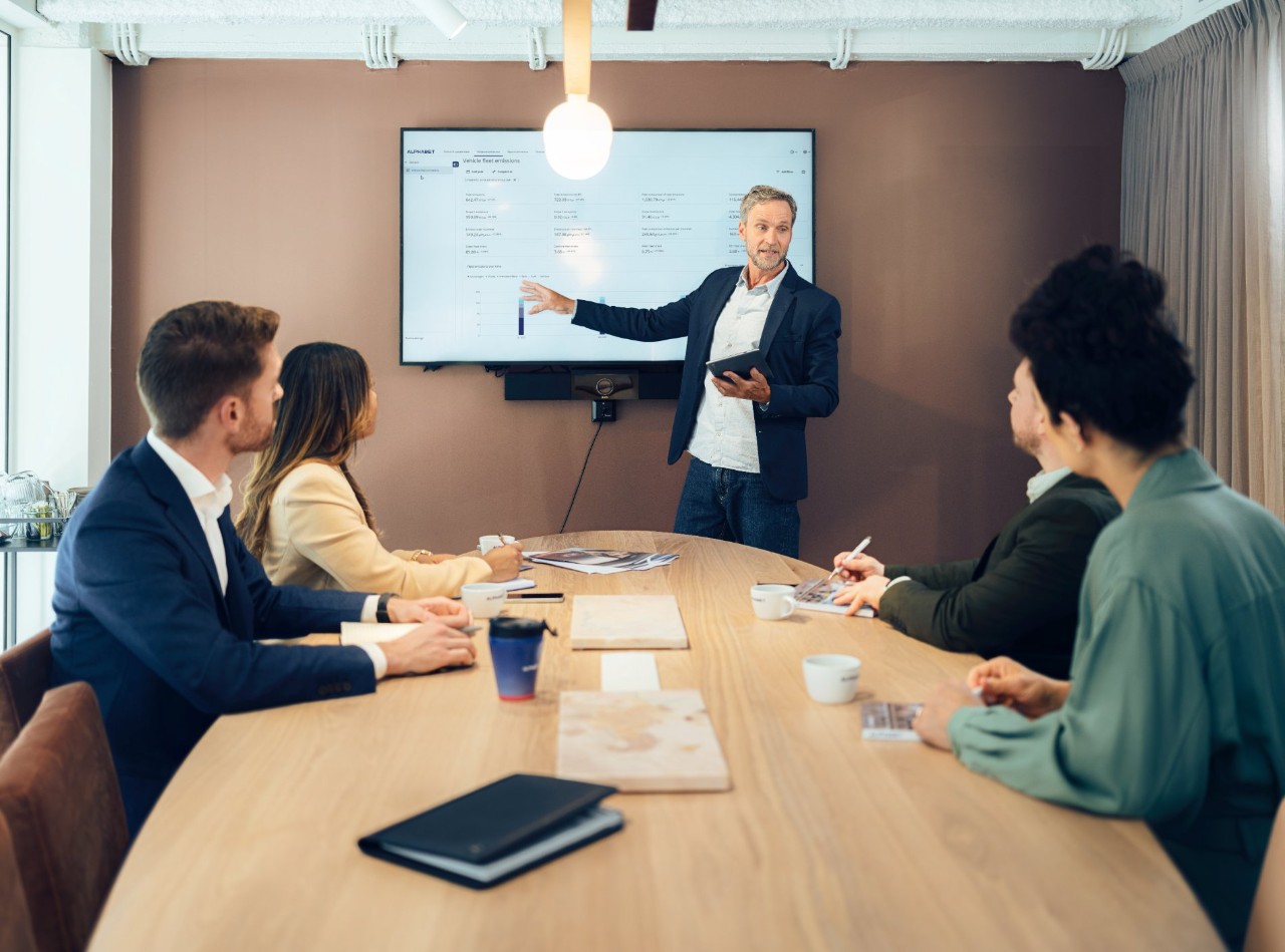Man stood up pointing at screen in front of a desk with 4 people sat down writing notes