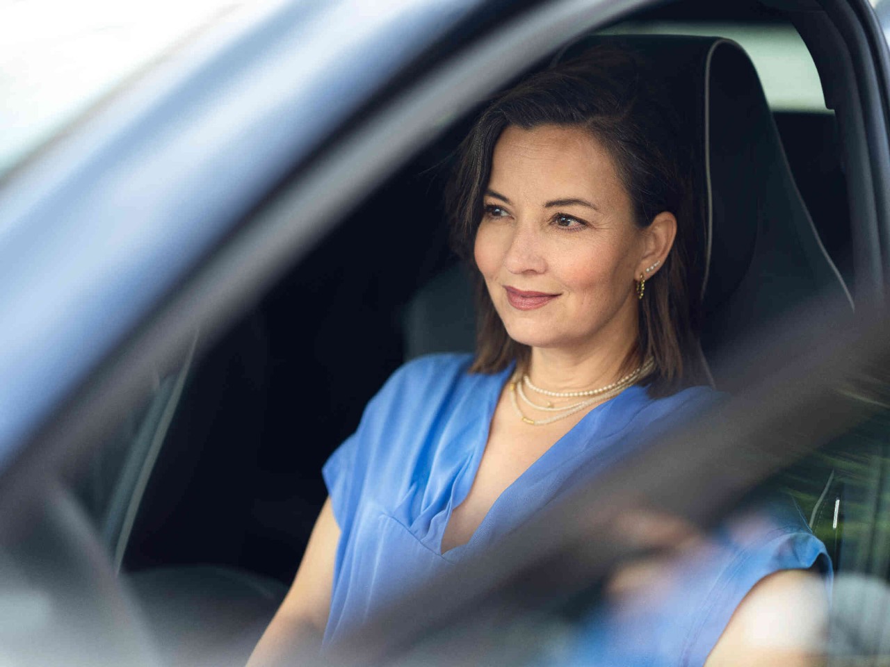 A woman sits in the driver’s seat of a car with the door open, looking intently ahead at the road.